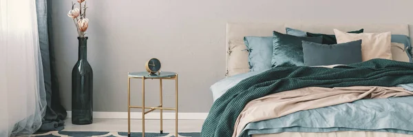 Fleurs en forme de bouteille vase à côté de l'horloge sur une table de chevet élégante avec des jambes dorées à côté du lit king size avec literie vert émeraude, bleu et beige — Photo