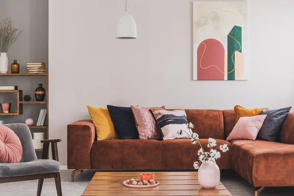 Real photo of a cozy, brown corner sofa with decorative cushions standing in a bright living room interior with an abstract painting — 스톡 사진