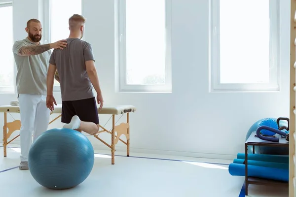 Jonge professionele fysiotherapeut die traint met een gewonde patiënt met een blauwe bal — Stockfoto