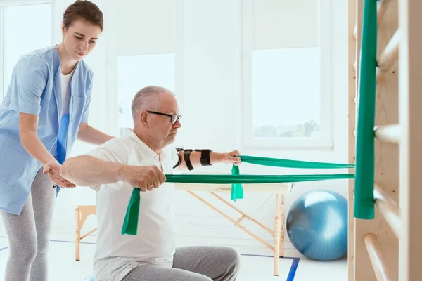 Starszy mężczyzna ćwiczy gimnastykę z młodą fizjoterapeutką — Zdjęcie stockowe