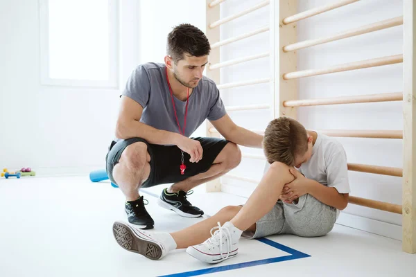 Joven entrenador ayuda al niño lesionado en el gimnasio de la escuela — Foto de Stock