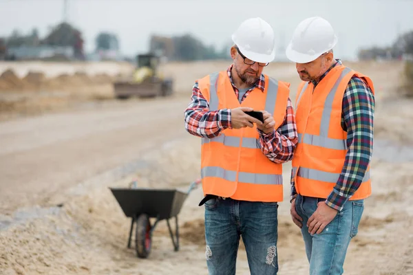 Two builders checking road construction plan on the phone — Stock Photo, Image