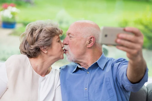 Bonito casal de idosos se beijando na frente de um telefone celular enquanto toma selfie — Fotografia de Stock