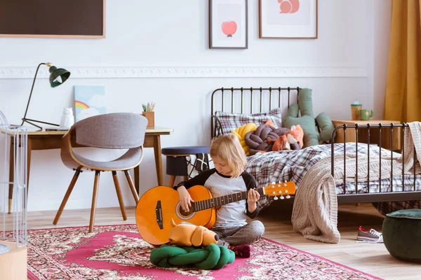 Linda chica rubia sentada en el suelo de su dormitorio de moda, aprendiendo a tocar una guitarra —  Fotos de Stock