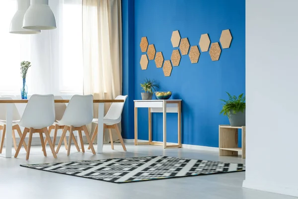 Korkwaben an der blauen Wand des trendigen Esszimmers mit gemustertem Teppich, Pflanzen und weißen Möbeln — Stockfoto