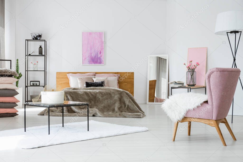 Trendy white bedroom with king size bed, industrial shelf and stylish pink armchair, abstract painting on empty wall