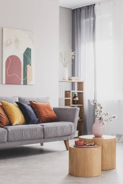 Pastel pembe vazodaki çiçekler ahşap sehpanın üzerinde renkli yastıklı gri İskandinav kanepesinin önünde.