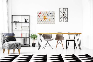 Gri koltukta yastık, geometrik sırt ve modaya uygun yemek odasında beyaz halı, duvarda saat ve poster ve yerde bitki.