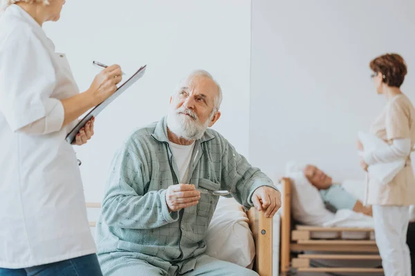 Bej Üniformalı Kıdemli Doktor Hastanede Yürürken Yaşlı Bir Hastayla Konuşuyor — Stok fotoğraf