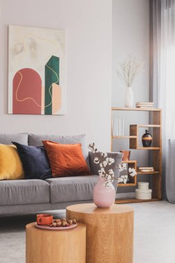 Pastel pembe vazodaki çiçekler ahşap sehpanın üzerinde renkli yastıklı gri İskandinav kanepesinin önünde.