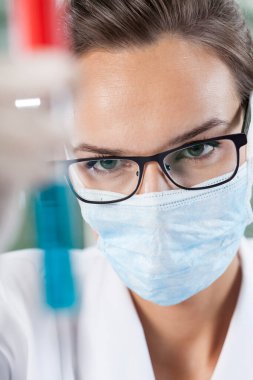 Cerrahi maskeli kadın bilim adamı mavi sıvıyla test tüpünü inceliyor.