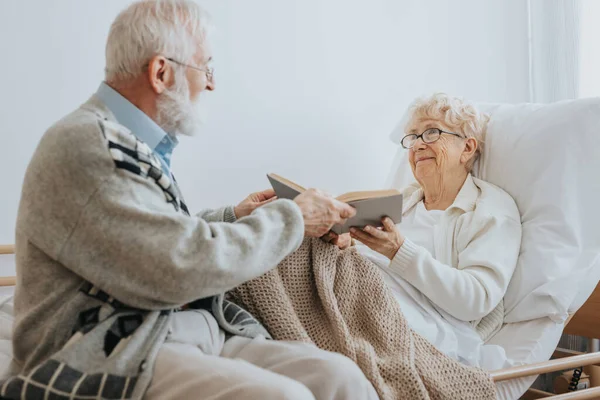 stock image Grey elderly man hands a book to a senior friend lying on a hospital bed