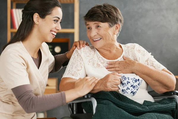 坐在轮椅上的积极的老年女士举着一杯草药茶 微笑着面对疗养院里漂亮的志愿者 — 图库照片