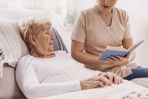 Hjälpsam Ung Vårdare Läser Bok För Äldre Kvinna Som Ligger — Stockfoto