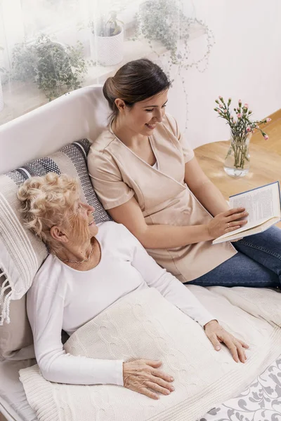 一位乐于助人的女性志愿者给躺在床上的老年妇女读一本书 — 图库照片