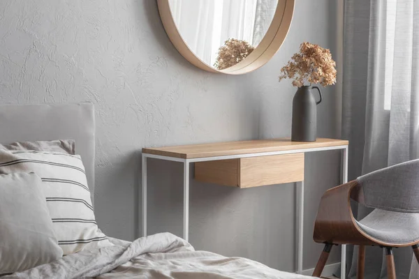 Elegant round mirror in wooden frame above fancy console table with flowers in vase in trendy bedroom interior with beige vase