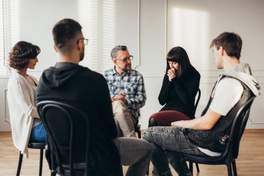 Farklı yaşlarda bir grup insan, profesyonel bir terapistle görüşürken bir çemberin içinde oturuyor.