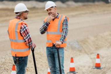 Arazinin ortasında turuncu yelekli ve koruyucu kasklı iki profesyonel yol inşaat işçisi var.