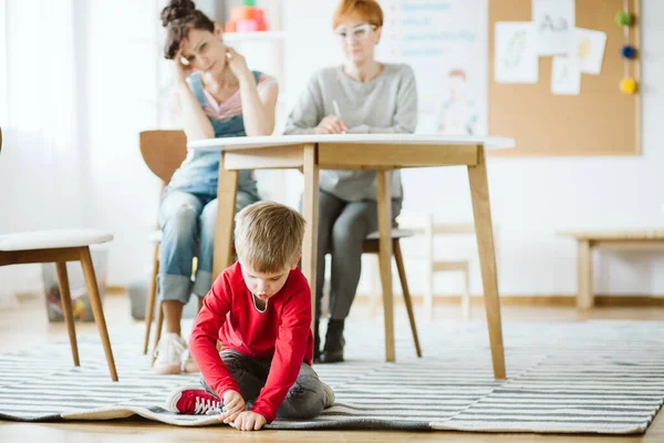 在与心理治疗师和他的母亲交谈时 年轻的自闭症男孩在地板上玩耍 — 图库照片