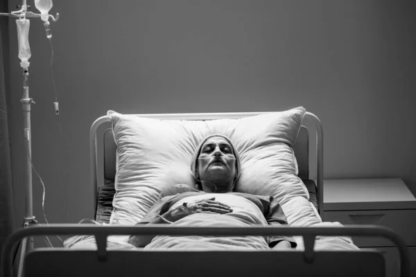 Suffering senior woman with cancer lying alone in hospital bed during chemotherapy, black and white photo