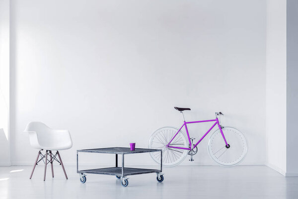 Purple bike, chair and metal coffee table in a minimalistic room interior. Real photo