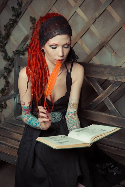 Tattooed girl with red dreadlocks, sitting on a bench, reading a book — Stock Photo, Image