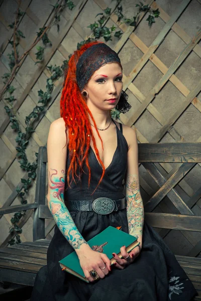 Dövmeli kız bir bankta oturan kırmızı dreadlocks bir kitap okuma