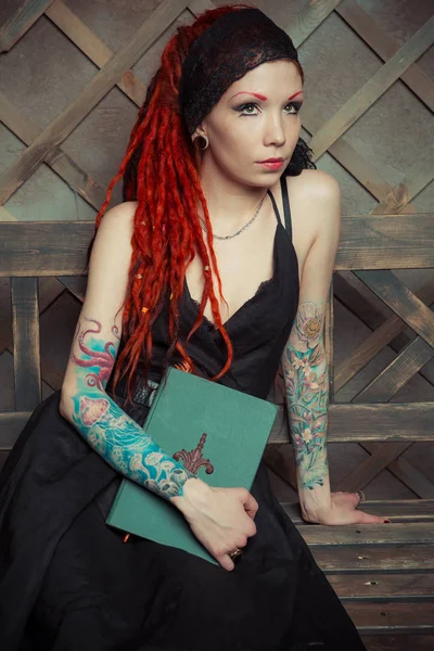 Chica tatuada con rastas rojas, sosteniendo un libro y una pluma, sentada en un banco, soñando con algo —  Fotos de Stock