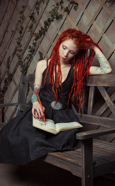 Tattooed girl with red dreadlocks, sitting on a bench, reading a book — Stock Photo, Image