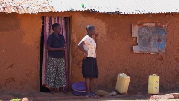 December, 2019. Afrika, Oeganda, dorp aan het Victoria meer. Twee vrouwen staan bij het huis, kleine jongen met een kind op zijn rug, rent naar hen toe en laat zien dat de toeristen hem snoep gaven — Stockvideo