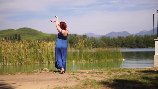 Чувственная рыжеволосая женщина в синем платье танцует на берегу озера, красиво расправляя руки аромат духов — стоковое видео