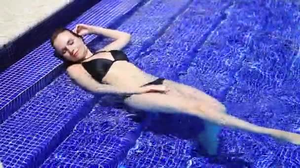 Belle fille en maillot de bain noir admire son corps, couché dans l'eau sur les marches de la piscine — Video