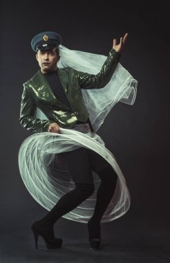 Koyu yeşil paltolu, şapkalı, duvak ve topuklu beyaz etekli büyüleyici bir karakteri canlandıran bir adam.