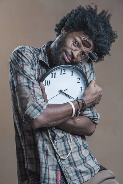 A curly-haired Dominican with open glasses on his glasses embraces a large wall clock and smiles