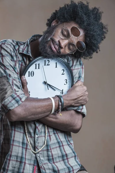 A curly-haired Dominican with open glasses on his glasses embraces a large wall clock and smiles