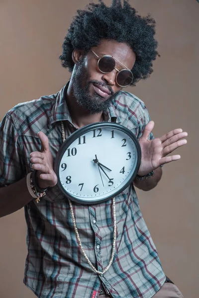 A curly-haired Dominican with open glasses on his glasses embraces a large wall clock and smiles