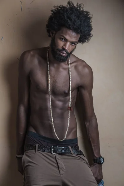 a dark-skinned, curly-haired guy with an athletic build , wearing long beads and his pants down