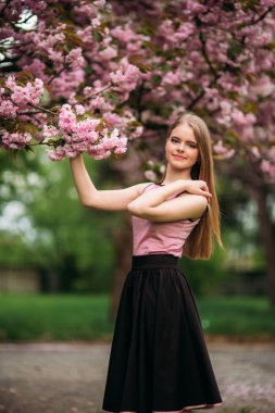 Sevimli kız pembe bluz ve siyah etek güzel çiçekli ağaçlar arka plan üzerinde fotoğrafçılar için poz veriyor. Bahar. Sakura.