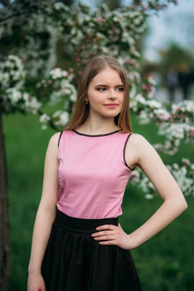 Charming girl in pink blouse and black skirt poses for photographers on the background of beautiful flowering trees. Spring. Sakura.