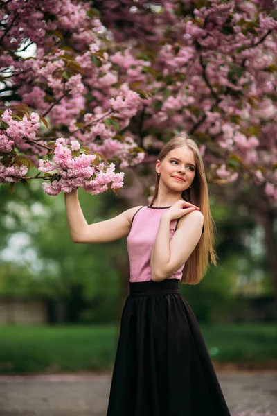 Uroczy dziewczyna w różowa bluzka i czarna spódnica stwarza dla fotografów na tle pięknych kwitnących drzew. Wiosną. Sakura. — Zdjęcie stockowe