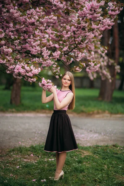 Charmig flicka i rosa blus och svart kjol poserar för fotograferna på bakgrunden av vackra blommande träd. Våren. Sakura. — Stockfoto