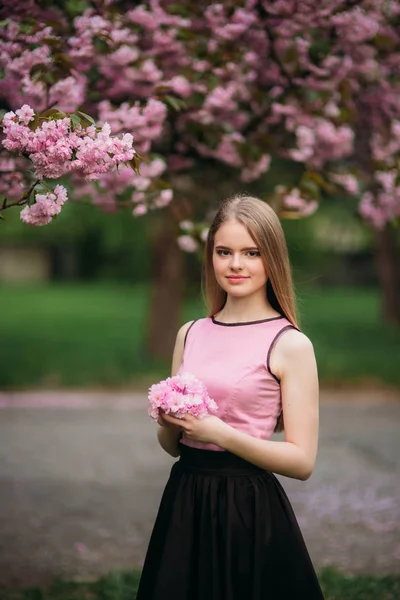 Charming girl in pink blouse and black skirt poses for photographers on the background of beautiful flowering trees. Spring. Sakura.