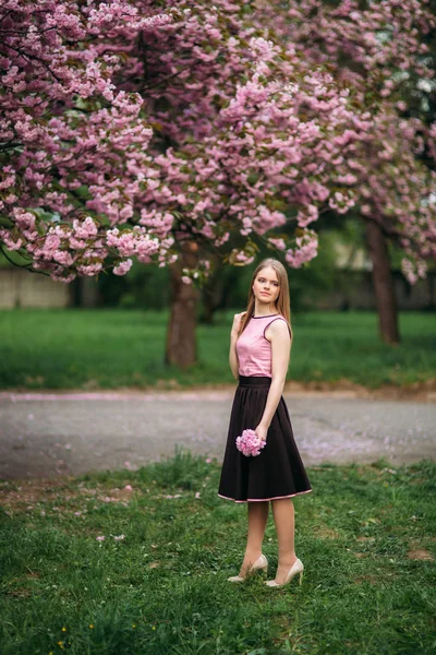 Charmig flicka i rosa blus och svart kjol poserar för fotograferna på bakgrunden av vackra blommande träd. Våren. Sakura. — Stockfoto