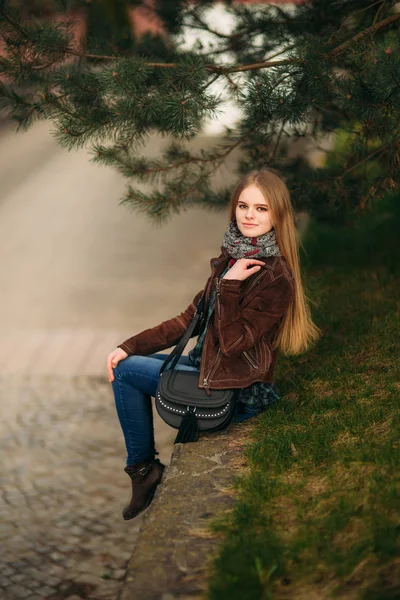 Una hermosa chica está caminando por el terraplén. Cabello rubio y chaqueta marrón. Primavera . —  Fotos de Stock