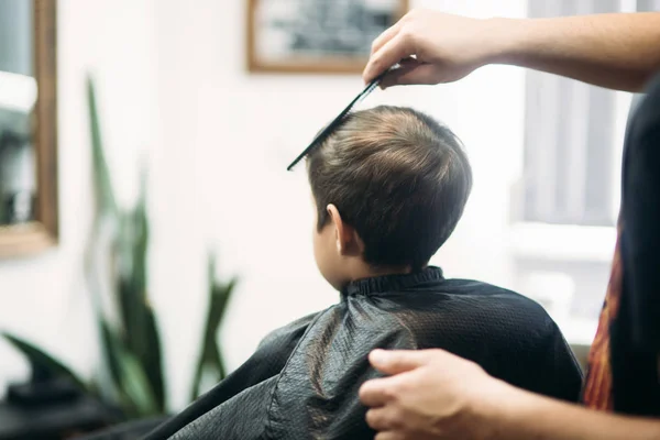 Pequeño chico consiguiendo corte de pelo por peluquero mientras está sentado en silla en la barbería . — Foto de Stock