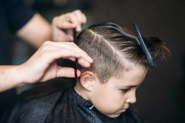 Pequeño chico consiguiendo corte de pelo por peluquero mientras está sentado en silla en la barbería . — Foto de Stock