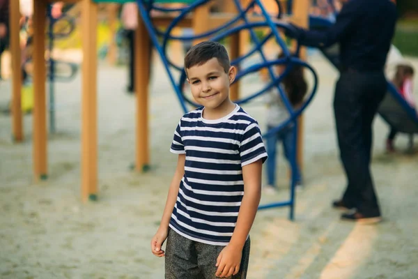 Un bambino con una maglietta a righe sta giocando nel parco giochi, oscillando su un'altalena.Primavera, tempo soleggiato . — Foto Stock