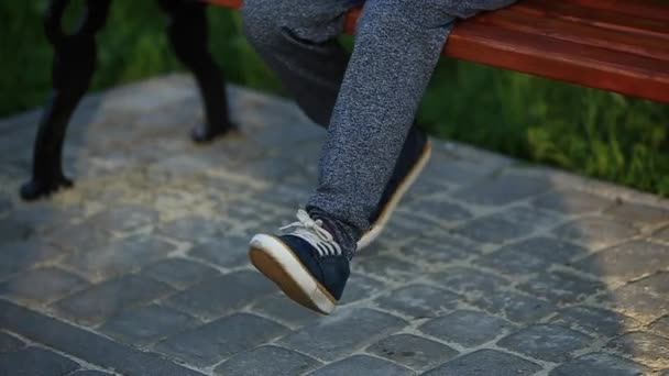 Un chico con pantalones y zapatillas sentado en un banco agitando las piernas. Puesta de sol de verano — Vídeos de Stock