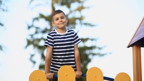 Un niño pequeño con una camiseta a rayas está jugando en el patio de recreo, columpio en un columpio.Pring, tiempo soleado . — Vídeo de stock