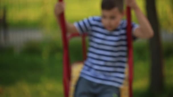 Маленький мальчик в полосатой футболке играет на детской площадке, качается на качелях. . — стоковое видео